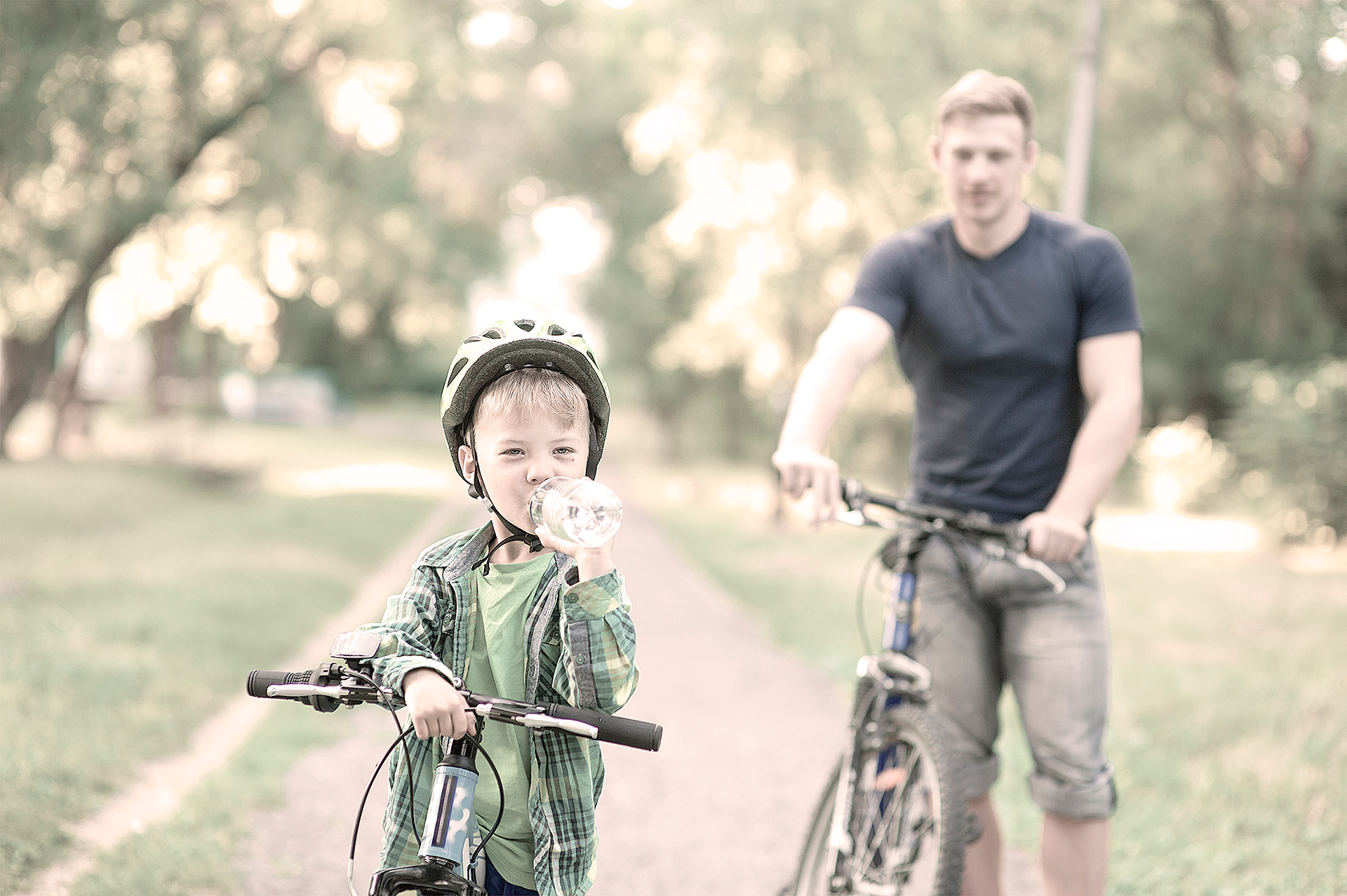 Dreng står med sin cykel, mens han drikker af en vandflaske. Far står i baggrunden med sin cykel og kigger på drengen
